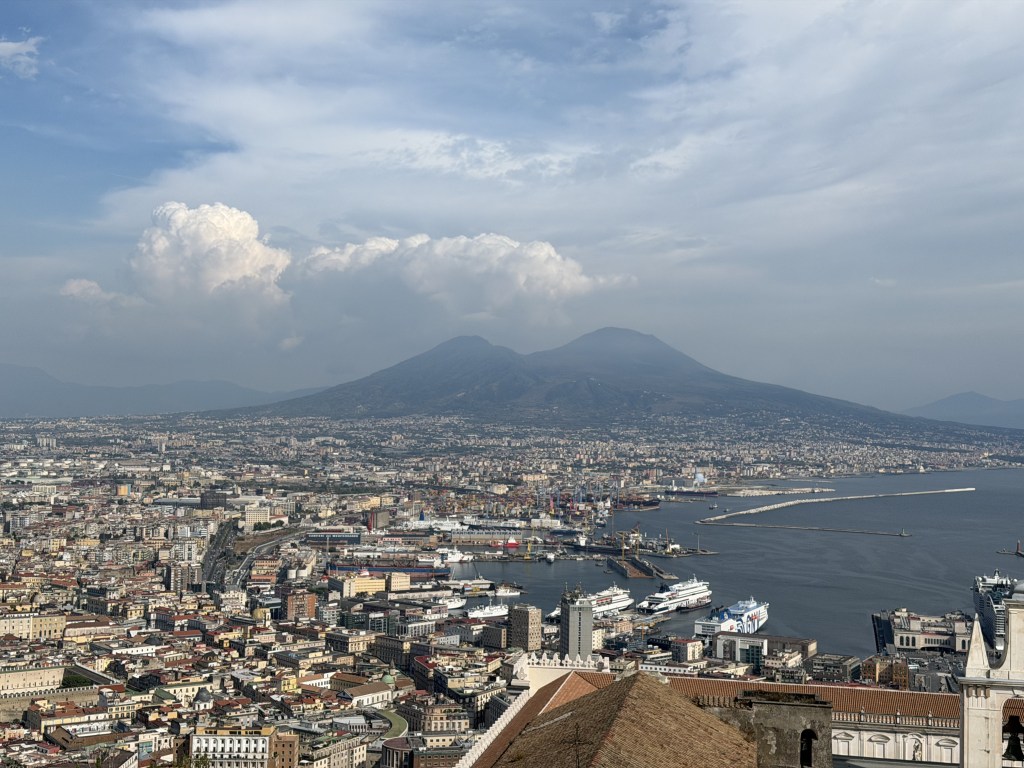A panoramic view of Naples, Italy, featuring the bustling cityscape, harbor, and Mount Vesuvius in the background under a partly cloudy sky.