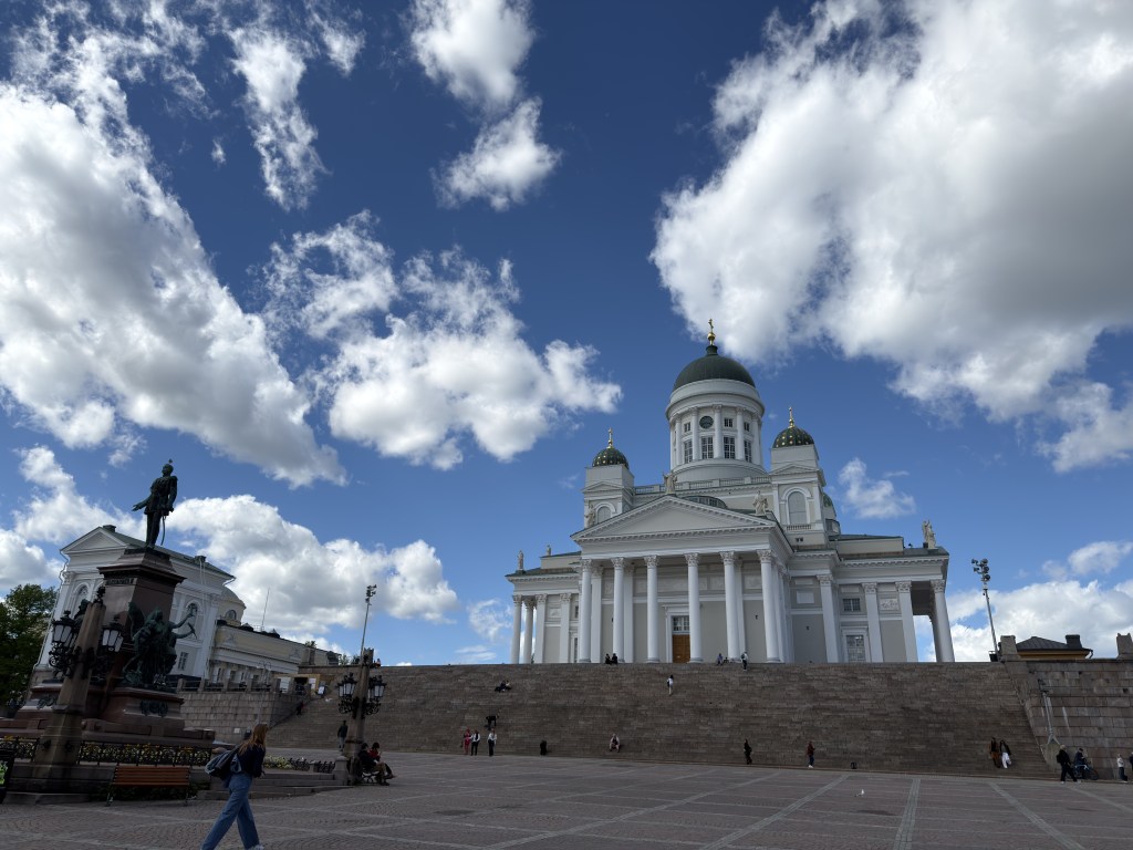 A view of the Helsinki Cathedral with a large set of stairs leading up to it, against a backdrop of cloudy blue sky. A statue is visible in the foreground, and several people are walking around in the square.